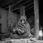 Na imagem, a jovem está no centro da cena, sentada no chão. Ela veste um manto com capuz e está rodeada por papéis e revistas abertas. À esquerda, há uma mala antiga aberta no chão; à direita, uma lamparina acesa ilumina suavemente o ambiente. O cenário parece ser um sótão ou espaço rústico, com paredes simples, cordas penduradas e objetos armazenados ao fundo.