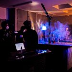 Cena de bastidores de gravação em estúdio: várias pessoas com máscaras operam câmeras e equipamentos de iluminação em um ambiente escuro, com fumaça azulada ao fundo. Sobre a bancada, há frascos de laboratório iluminados, e ao fundo vê-se um esqueleto humano em pé.