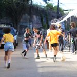 Crianças jogam futebol em uma rua enquanto adultos e equipe técnica acompanham a cena, com câmeras e refletores montados ao fundo.