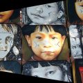 Fotografia mostra uma exposição de pinturas realistas e estilizadas de rostos de crianças indígenas com pinturas faciais tradicionais, dispostas lado a lado em painéis.
