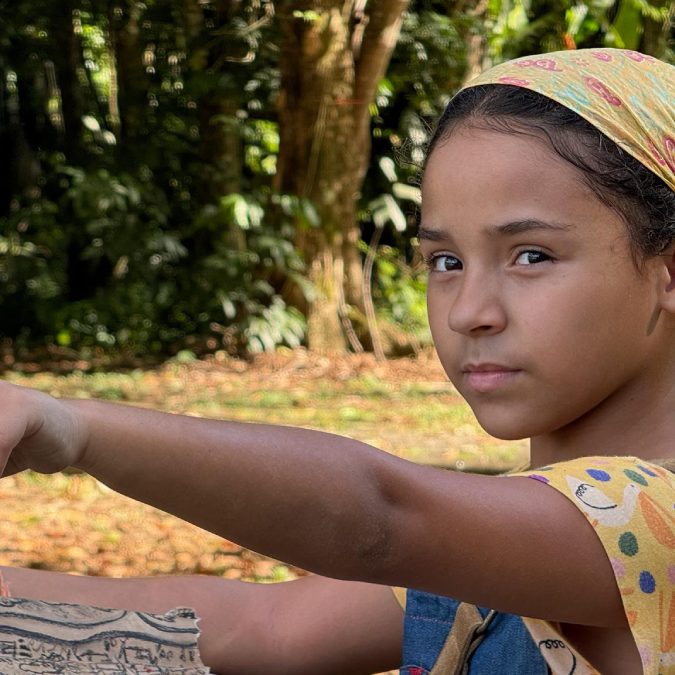 Menina com lenço amarelo estampado na cabeça segura um mapa antigo e aponta para frente com expressão séria, em meio a uma área arborizada.