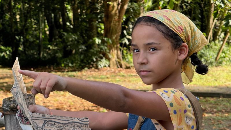 Menina com lenço amarelo estampado na cabeça segura um mapa antigo e aponta para frente com expressão séria, em meio a uma área arborizada.