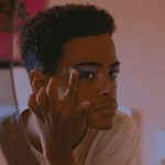 Jovem olha para o espelho enquanto aplica sombra azul na pálpebra com o dedo, em um ambiente iluminado por luz suave e avermelhada, com fotos coladas na parede ao lado.