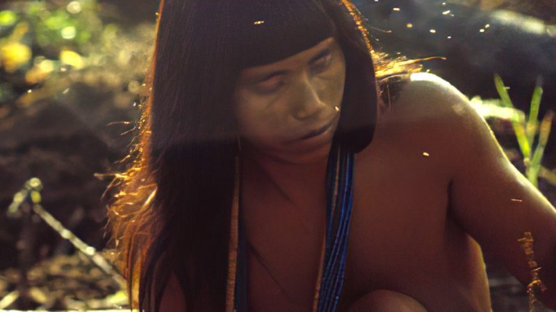 Pessoa indígena com pintura facial clara e colares azuis aparece em cena iluminada pela luz do sol, cercada por elementos da natureza.