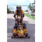 Duas crianças usando roupas amarelas estampadas, capacetes e joelheiras descem uma rua em um carrinho de rolimã decorado como cabeça de animal, sorrindo enquanto brincam.