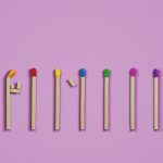 Sete fósforos coloridos estão alinhados sobre fundo lilás, com cabeças em laranja, vermelho, amarelo, azul, verde, rosa e roxo. O primeiro fósforo está curvado com pequenos pedaços de madeira colados, sugerindo a forma de uma figura humana.