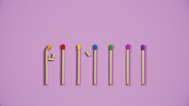 Sete fósforos coloridos estão alinhados sobre fundo lilás, com cabeças em laranja, vermelho, amarelo, azul, verde, rosa e roxo. O primeiro fósforo está curvado com pequenos pedaços de madeira colados, sugerindo a forma de uma figura humana.