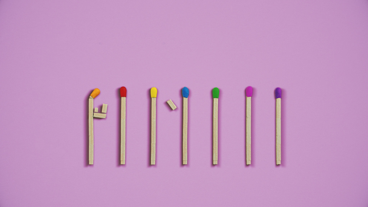 Sete fósforos coloridos estão alinhados sobre fundo lilás, com cabeças em laranja, vermelho, amarelo, azul, verde, rosa e roxo. O primeiro fósforo está curvado com pequenos pedaços de madeira colados, sugerindo a forma de uma figura humana.