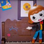 Boneca de animação com cabelos castanhos curtos, roupas coloridas e expressão sorridente está em pé diante de um piano de madeira decorado com desenhos. Ao fundo, prateleiras com brinquedos e livros, além de quadros e desenhos infantis na parede. No canto superior esquerdo aparece o logotipo “Sol em Do Ré Mi”.