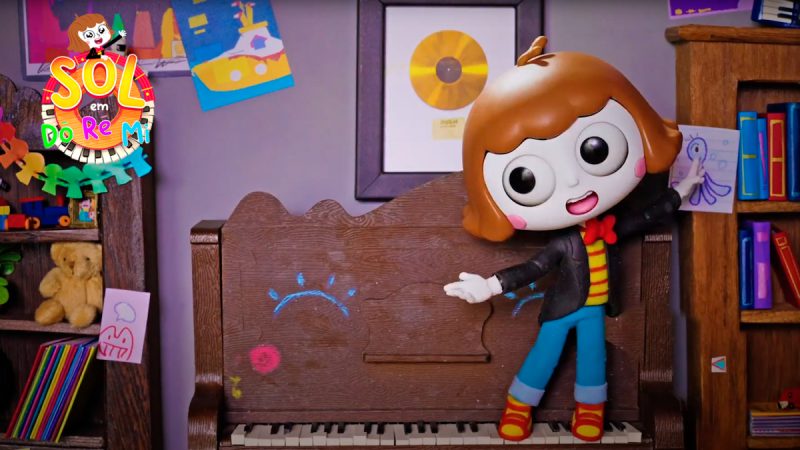 Boneca de animação com cabelos castanhos curtos, roupas coloridas e expressão sorridente está em pé diante de um piano de madeira decorado com desenhos. Ao fundo, prateleiras com brinquedos e livros, além de quadros e desenhos infantis na parede. No canto superior esquerdo aparece o logotipo “Sol em Do Ré Mi”.