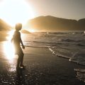 Silhueta de uma criança caminha à beira-mar durante o pôr do sol, com ondas quebrando suavemente e montanhas ao fundo.