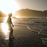 Silhueta de uma criança caminha à beira-mar durante o pôr do sol, com ondas quebrando suavemente e montanhas ao fundo.