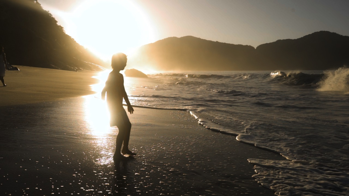 Silhueta de uma criança caminha à beira-mar durante o pôr do sol, com ondas quebrando suavemente e montanhas ao fundo.