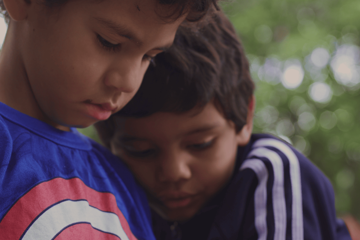 Dois meninos estão próximos, um vestindo camiseta azul com estampa circular vermelha e branca, e o outro usando agasalho azul-marinho com listras brancas, ambos com expressão concentrada.