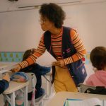 Professora entrega uma folha para uma aluna em sala de aula, enquanto outras crianças sentadas em suas carteiras observam e escrevem.