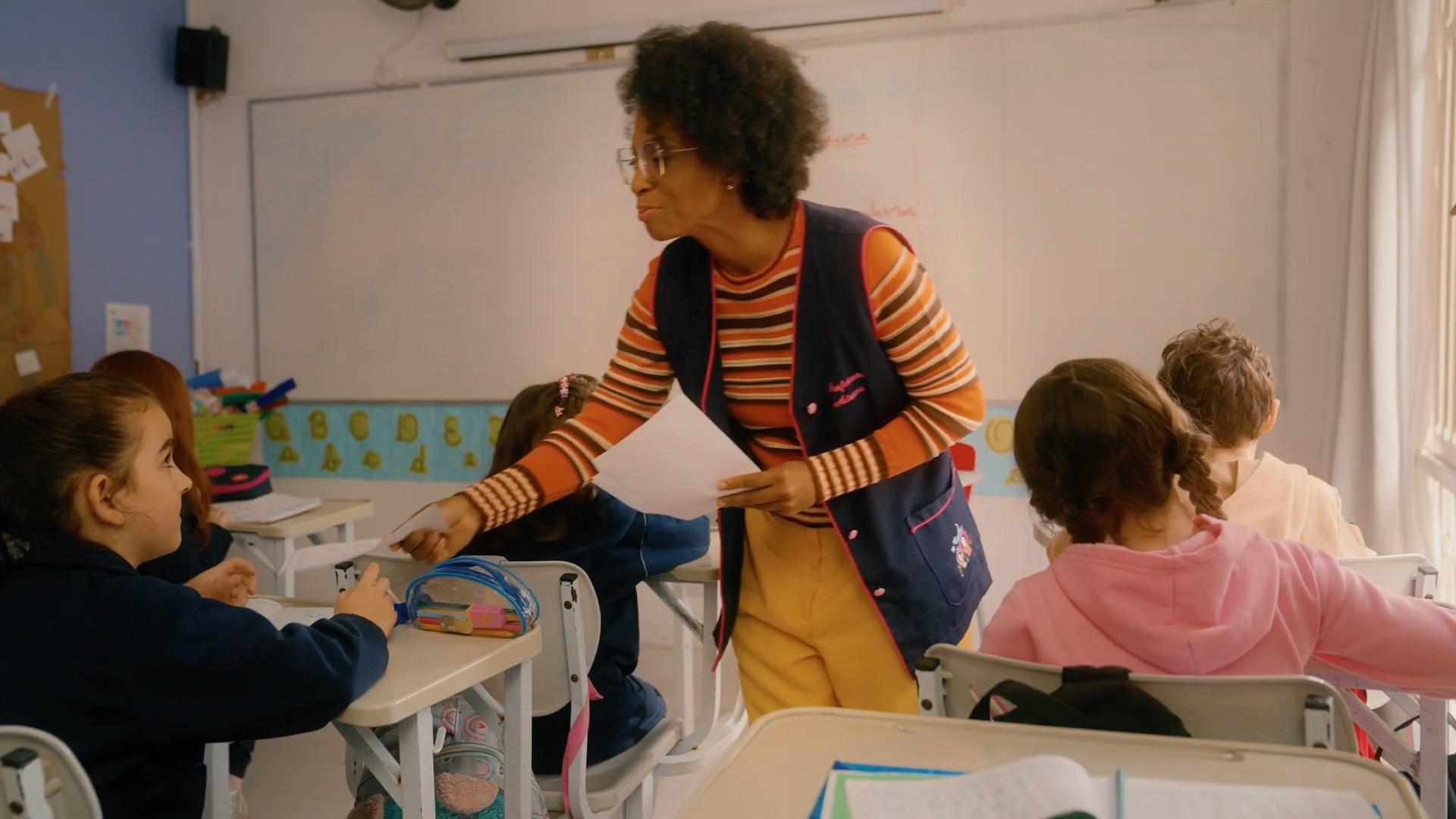 Professora entrega uma folha para uma aluna em sala de aula, enquanto outras crianças sentadas em suas carteiras observam e escrevem.