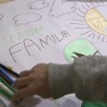 Desenho infantil em uma folha de papel com o título “Minha Família”, cercado por nuvens e um sol colorido em dourado. Ao lado estão lápis e canetas coloridas, enquanto uma mão de criança segura uma das canetas.