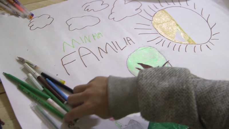 Desenho infantil em uma folha de papel com o título “Minha Família”, cercado por nuvens e um sol colorido em dourado. Ao lado estão lápis e canetas coloridas, enquanto uma mão de criança segura uma das canetas.