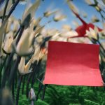 A imagem mostra um pedaço de papel vermelho em destaque, colocado entre flores brancas feitas de origami sobre um fundo verde que lembra fios de lã, com o céu azul ao fundo.