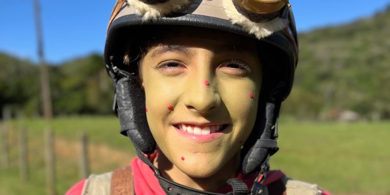 Criança com rosto pintado de verde, usando capacete com luz e óculos grandes no topo, sorrindo ao ar livre em dia ensolarado.