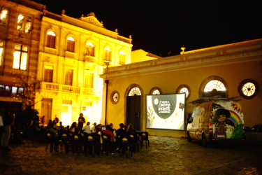 Projeção de cinema ao ar livre à noite, com pessoas sentadas em cadeiras diante de telão iluminado em pátio histórico.