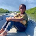 Menino sentado descalço na proa de um barco metálico, com expressão séria, cercado por águas calmas e vegetação verde.