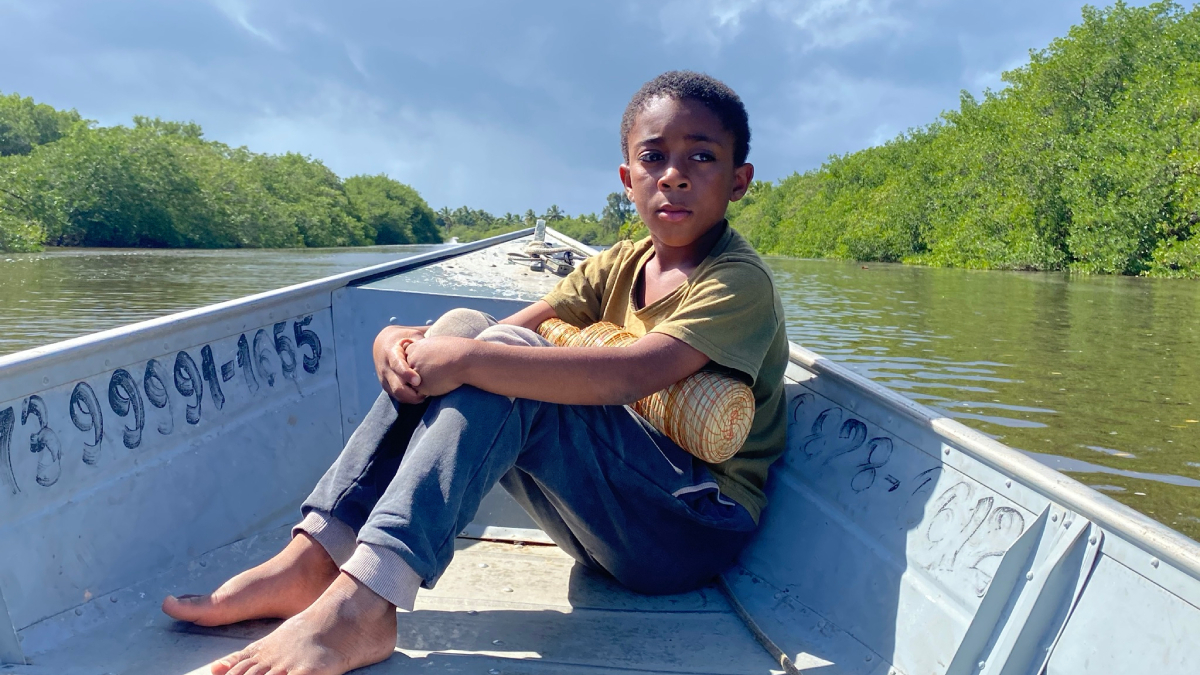 Menino sentado descalço na proa de um barco metálico, com expressão séria, cercado por águas calmas e vegetação verde.