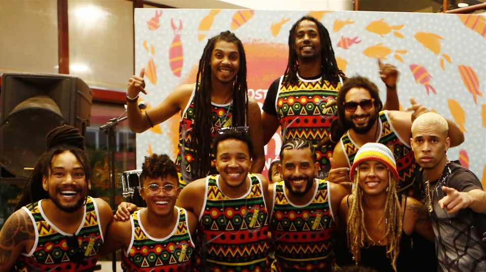 Grupo Africatarina Grupo de músicos sorridentes posa em frente a um painel colorido, usando camisetas iguais com estampas geométricas vibrantes.