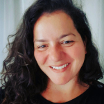 Mulher sorridente de cabelos longos e castanhos, em frente a uma cortina clara.