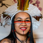 Mulher sorridente com cocar de penas coloridas e pintura indígena no rosto.