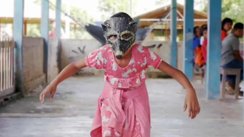 Fotografia; criança com máscara avança com braços estendidos, de vestido rosa, em pátio coberto; ao fundo, pessoas sentadas.