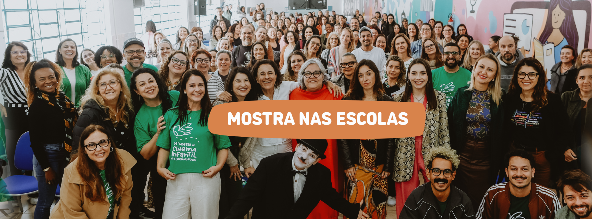 Um grande grupo de pessoas, em sua maioria educadores e organizadores, sorri reunido em auditório colorido, com a faixa central em laranja trazendo o texto “Mostra nas Escolas”.
