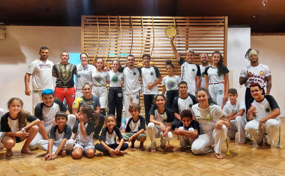 Arte do Cativeiro Capoeira Grupo diverso de crianças, jovens e adultos posa sorridente em roda de capoeira, alguns de uniforme branco e outros com camisetas pretas e brancas.