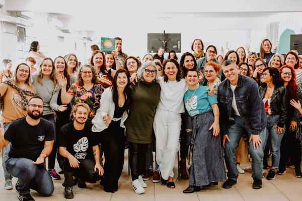 Encontro com professores na Mostra de 2024 Grupo grande de pessoas posando juntas em ambiente interno claro, muitas sorrindo e olhando para a câmera, transmitindo clima de confraternização e união.