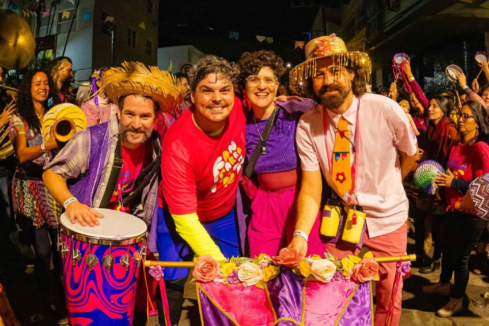 Fanfarra da Ponte Grupo sorridente em festa junina noturna, com roupas coloridas, chapéus de palha e instrumentos musicais.