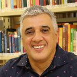 Homem sorridente de cabelos curtos grisalhos, em frente a estante de livros.