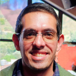 Homem sorridente de óculos e cabelos curtos castanhos escuros, com janela iluminada ao fundo.