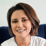 Mulher sorridente de cabelos curtos e castanhos, vestindo blusa clara, sentada em ambiente interno.