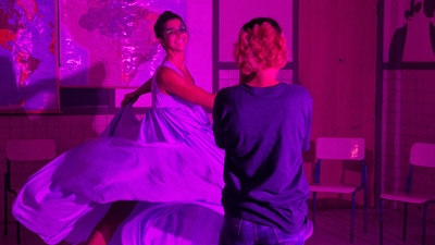 Mulher de vestido esvoaçante dança sorridente sob luz roxa intensa, enquanto outra pessoa a observa em ambiente com mapas na parede e cadeiras escolares.