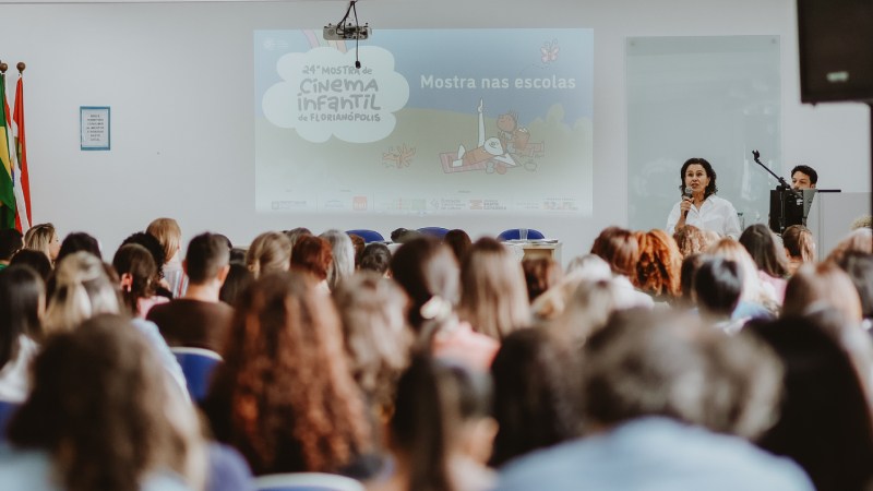 Auditório cheio de pessoas assistindo a uma apresentação, com tela projetada que mostra o texto “vigésima quarta Mostra de Cinema Infantil de Florianópolis – Mostra nas escolas”.