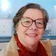 Maria Helena Michels Mulher madura sorridente, de óculos grandes, casaco bege e cachecol vermelho, em ambiente interno iluminado.