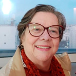 Mulher madura sorridente, de óculos grandes, casaco bege e cachecol vermelho, em ambiente interno iluminado.