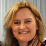 Mulher sorridente de cabelos loiros, usando lenço colorido no pescoço, com fundo de cortina clara.