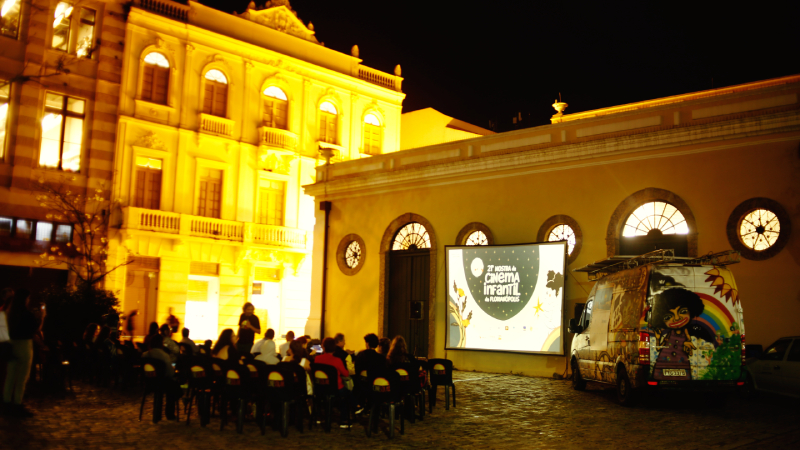 Projeção de cinema ao ar livre à noite, com pessoas sentadas em cadeiras diante de telão iluminado em pátio histórico.