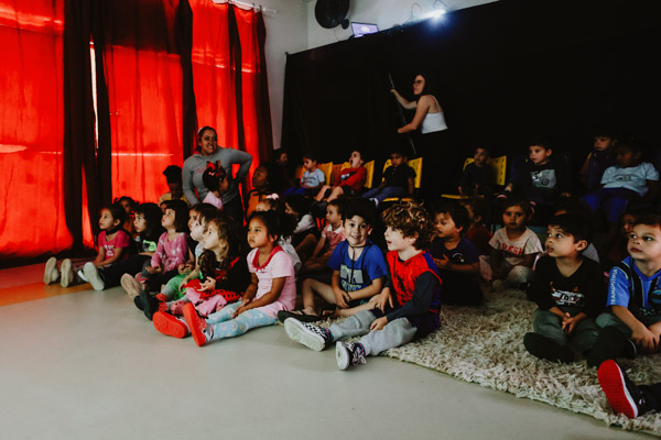 Grupo de crianças sentadas no chão de uma sala escura com cortinas vermelhas assistem atentas a uma projeção.