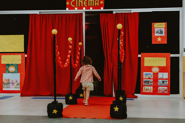 Criança de costas caminha sobre tapete vermelho em direção a cortinas vermelhas com placa “Cinema” acima da entrada decorada.