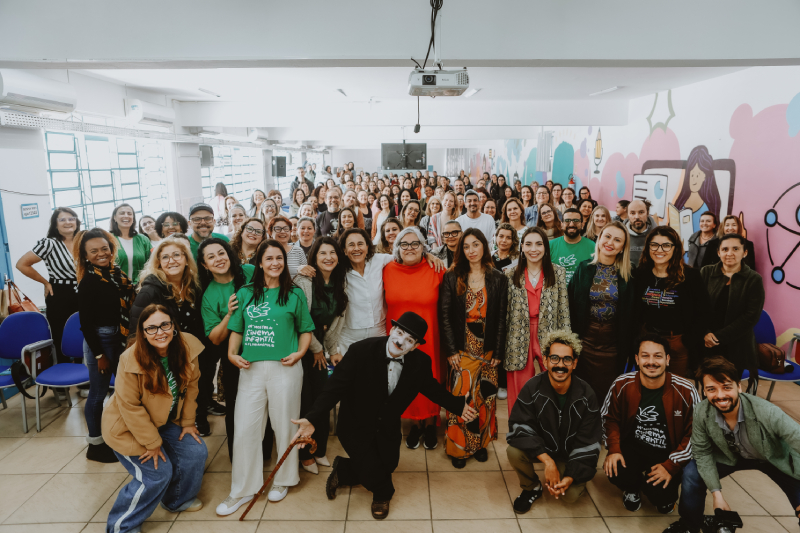 Mais de cem pessoas reunidas em um auditório, sorrindo para a foto, com destaque para um grupo na frente, incluindo um artista caracterizado como Charlie Chaplin.