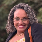 Retrato: mulher sorridente, cabelos cacheados, óculos e blazer preto; fundo verde desfocado.