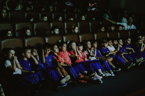 Grupo de crianças uniformizadas de azul e vermelho sentadas lado a lado em poltronas de cinema.