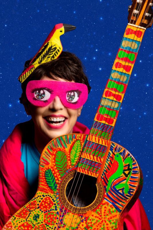 Pessoa sorridente com máscara rosa nos olhos, chapéu em forma de pássaro e violão colorido pintado à mão.